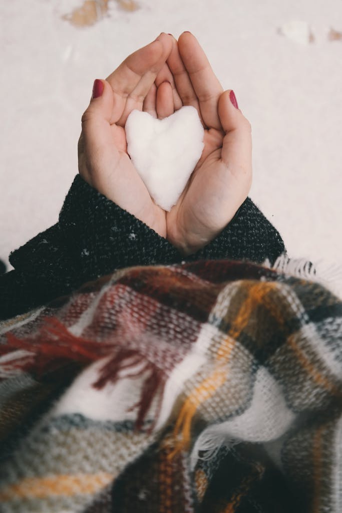 Close-up of hands holding a heart-shaped snow piece, wrapped in winter clothing.